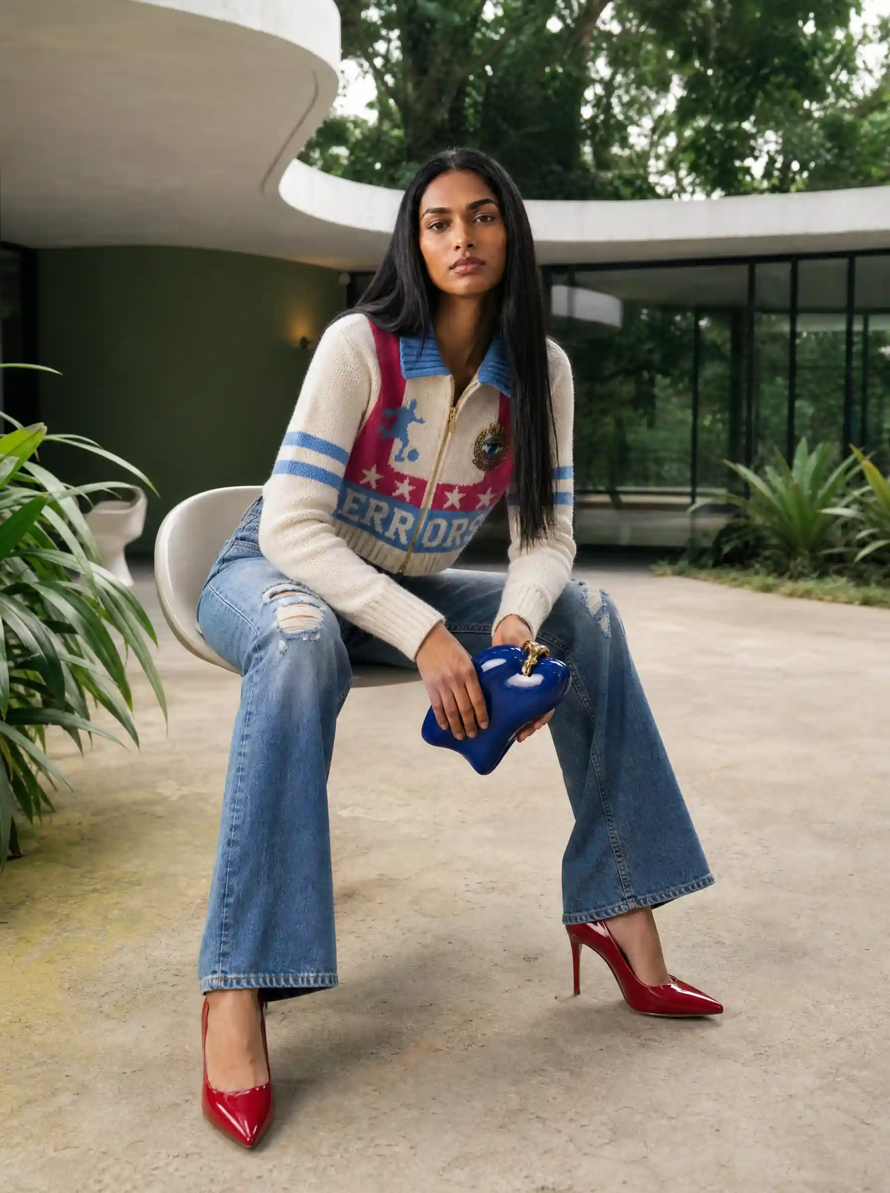 Dark-haired model in a varsity cardigan and flared jeans with red heels, seated holding a blue clutch outside a modern villa