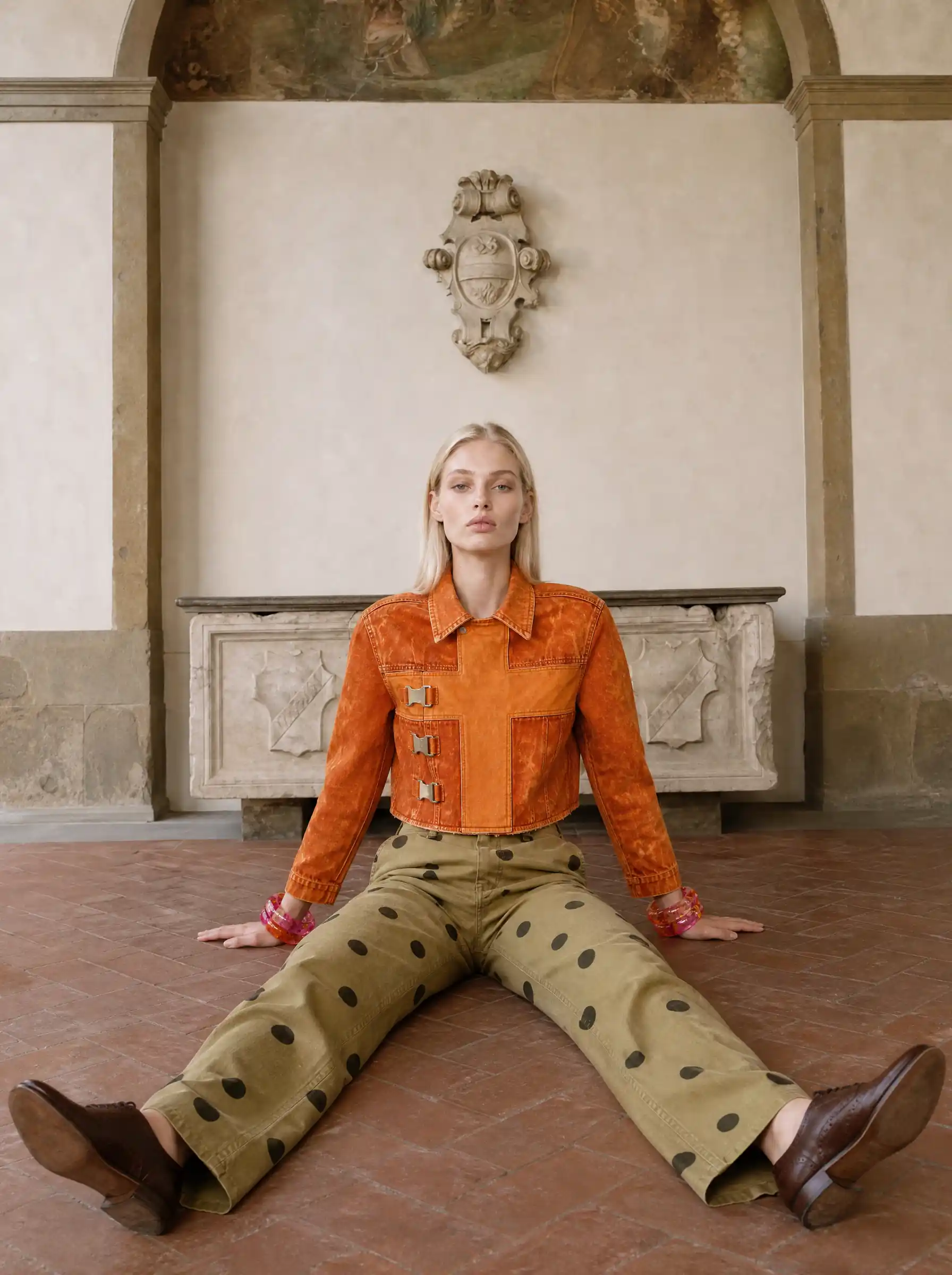 Blonde model in an orange suede jacket and olive polka-dot trousers, seated on a tiled floor in a frescoed old room