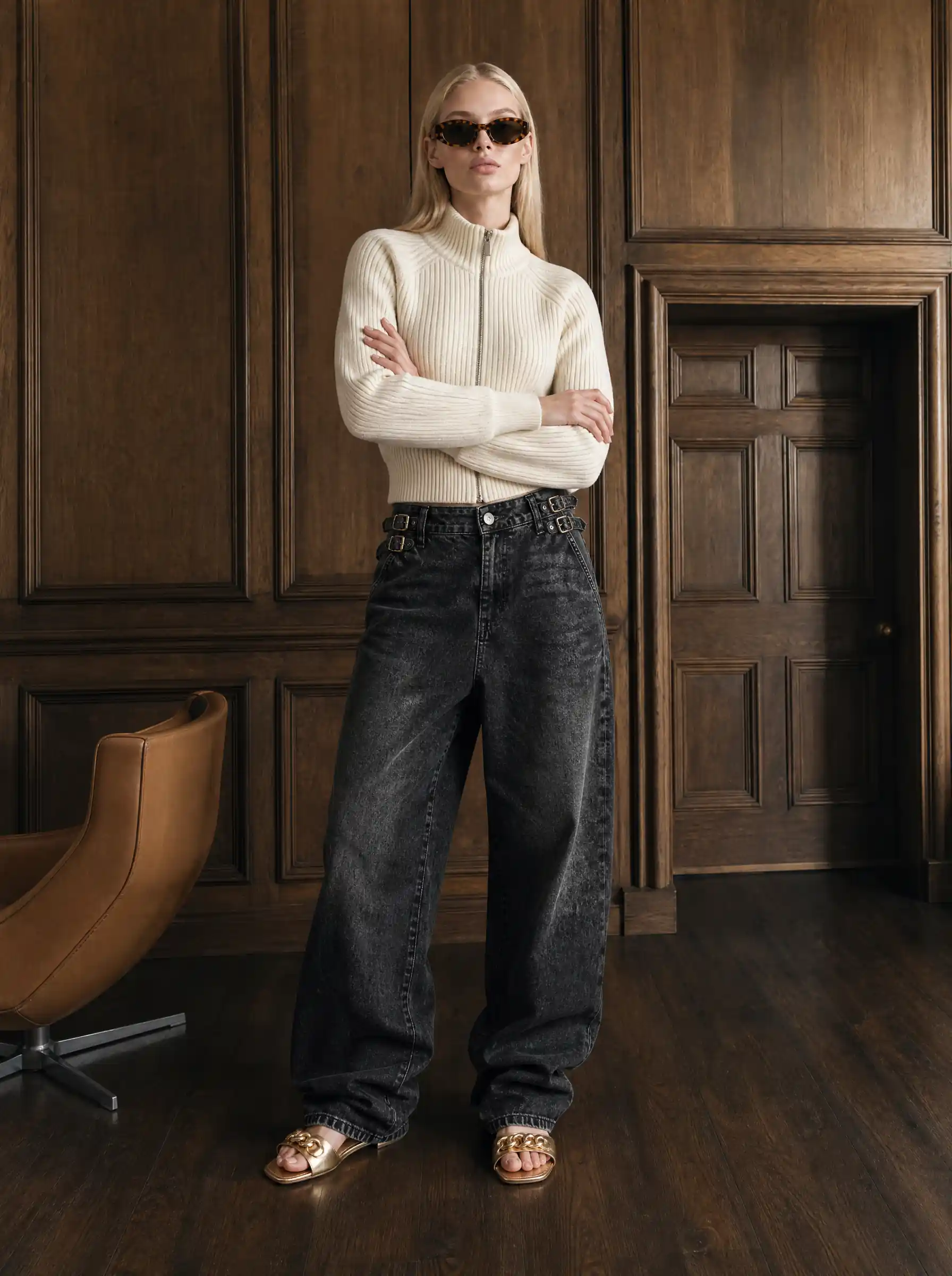 Blonde model in a cream zip sweater, baggy black jeans and gold sandals, standing in a wood-panelled room