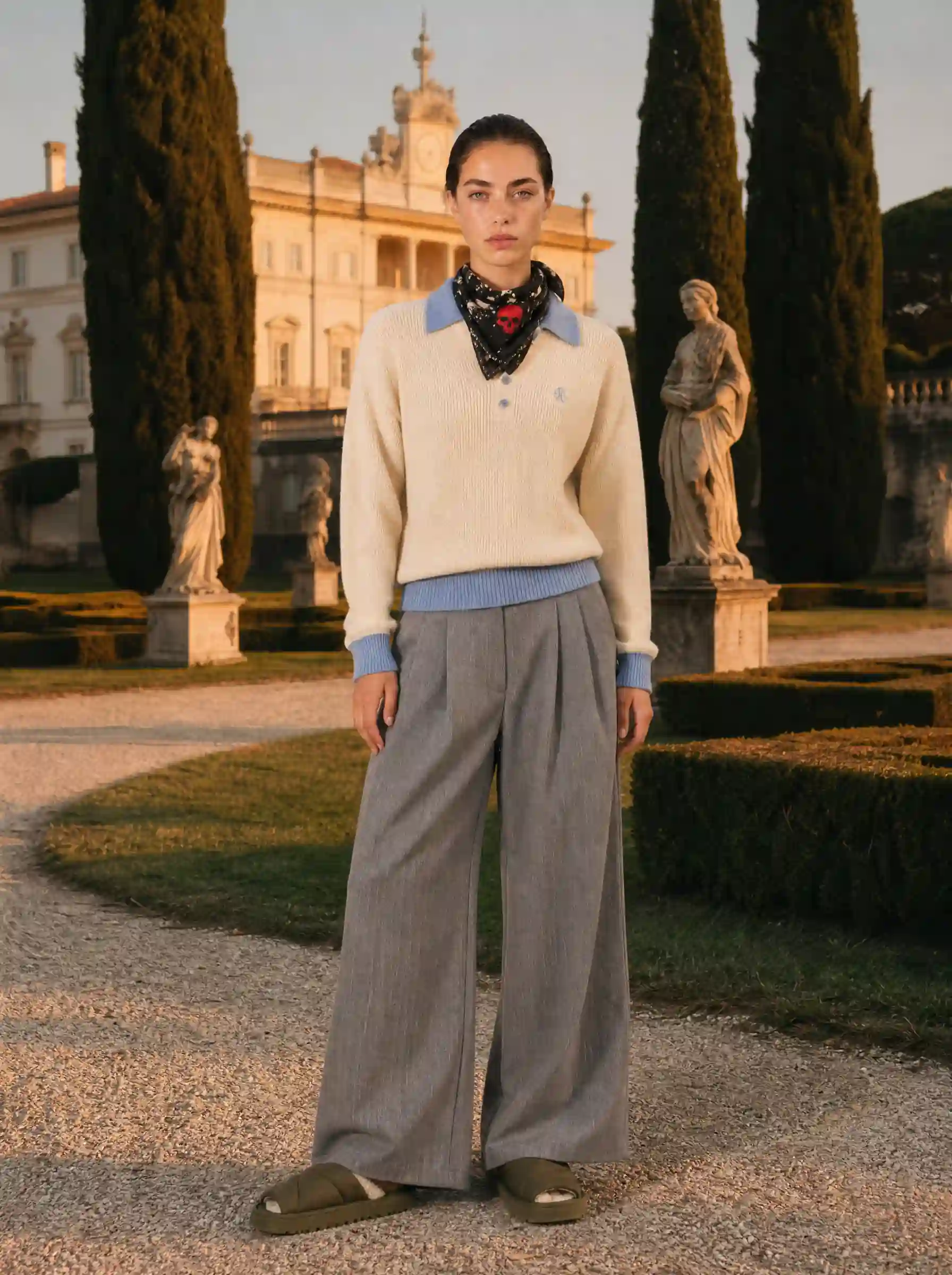 Model in a cream sweater with neck scarf and grey wide-leg trousers, in a formal garden with cypress trees and statues