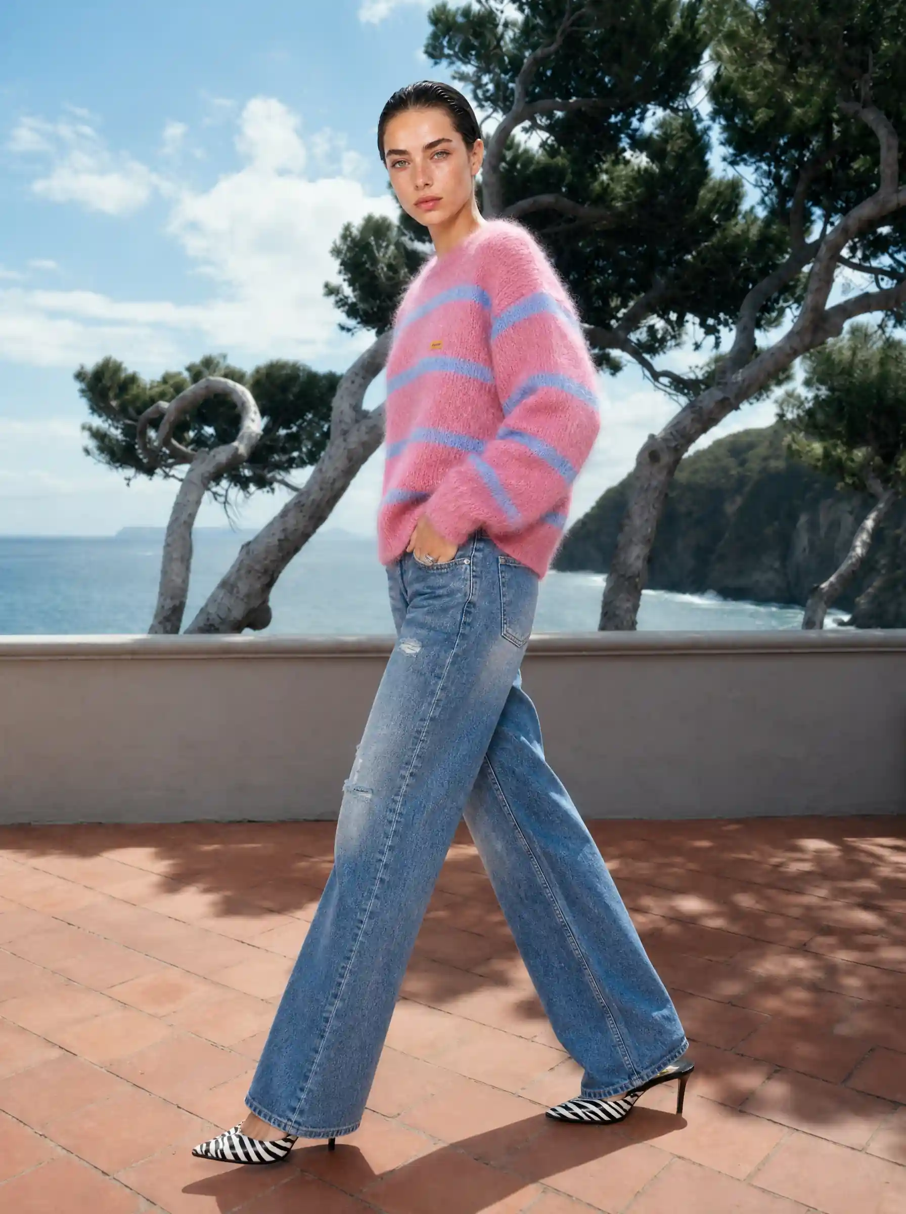 Model in a pink striped mohair sweater and wide-leg jeans, walking a sunlit coastal terrace above the sea