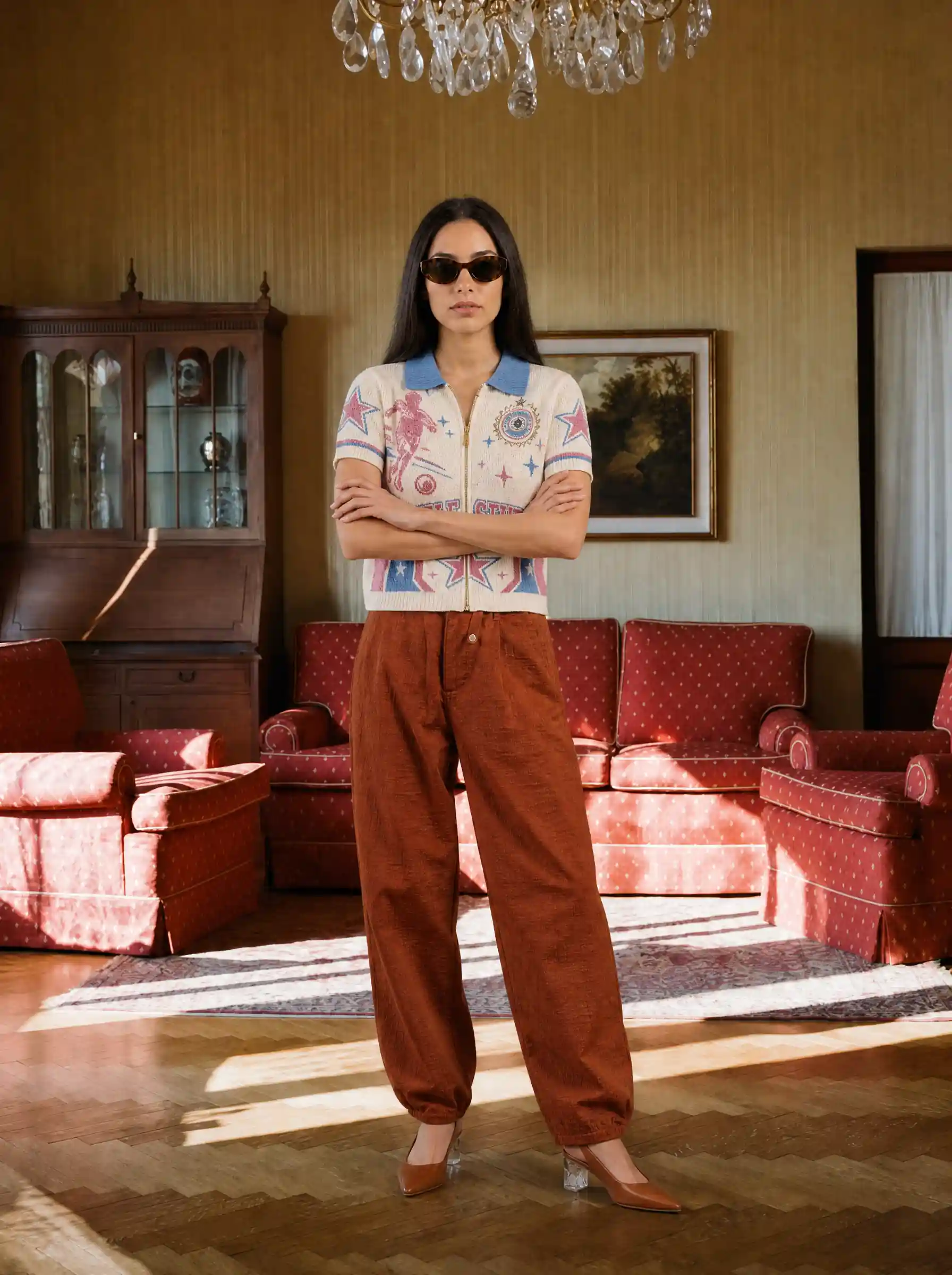 Dark-haired model in a printed zip polo and rust corduroy trousers, arms crossed in a vintage sitting room with red sofas