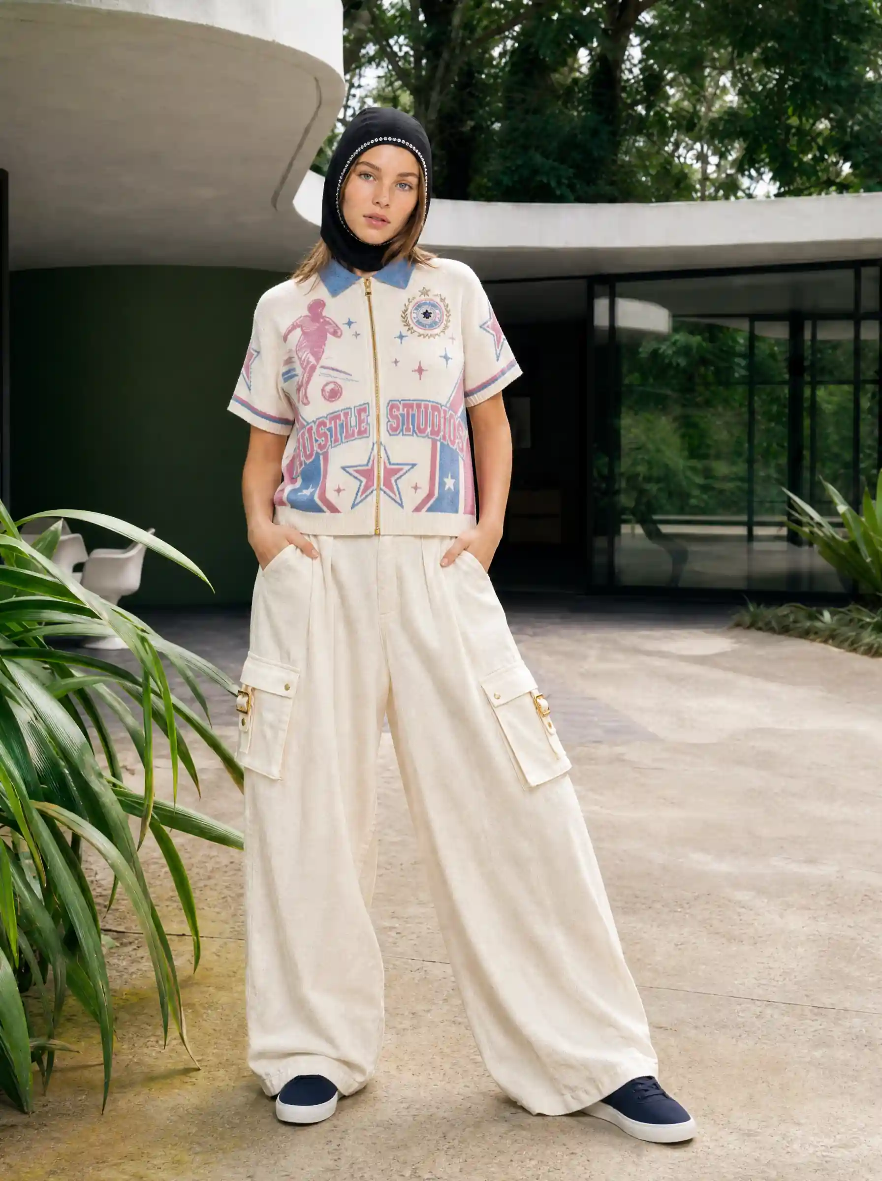 Model in a printed zip polo, cream cargo trousers and a knit hood, standing in a plant-filled modern courtyard