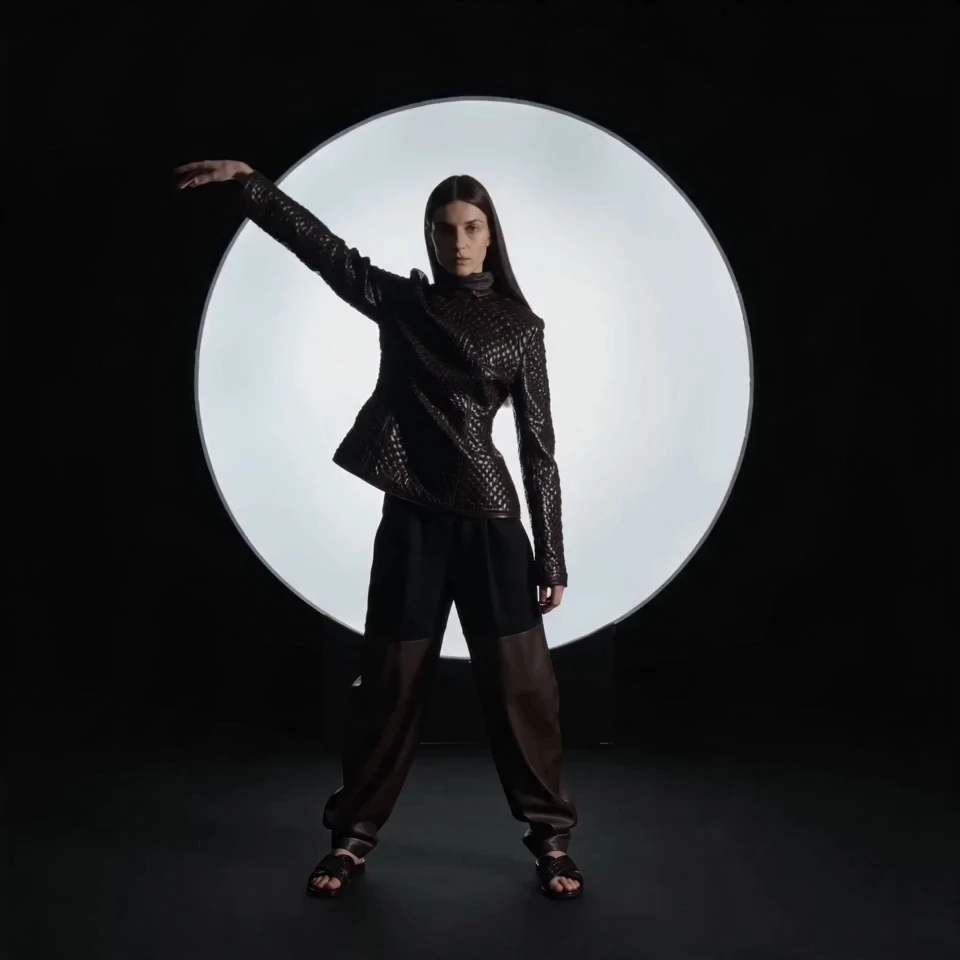Model in a textured black jacket posing with one arm raised, silhouetted against a large glowing white light