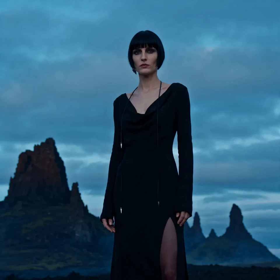 Model with a dark bob in a black cowl-neck gown, standing before rugged mountain peaks under a stormy blue sky