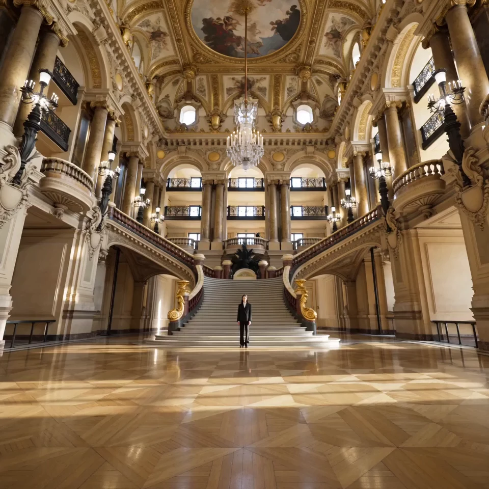 Lone figure in black standing in an opulent gilded palace hall with grand staircases and a crystal chandelier